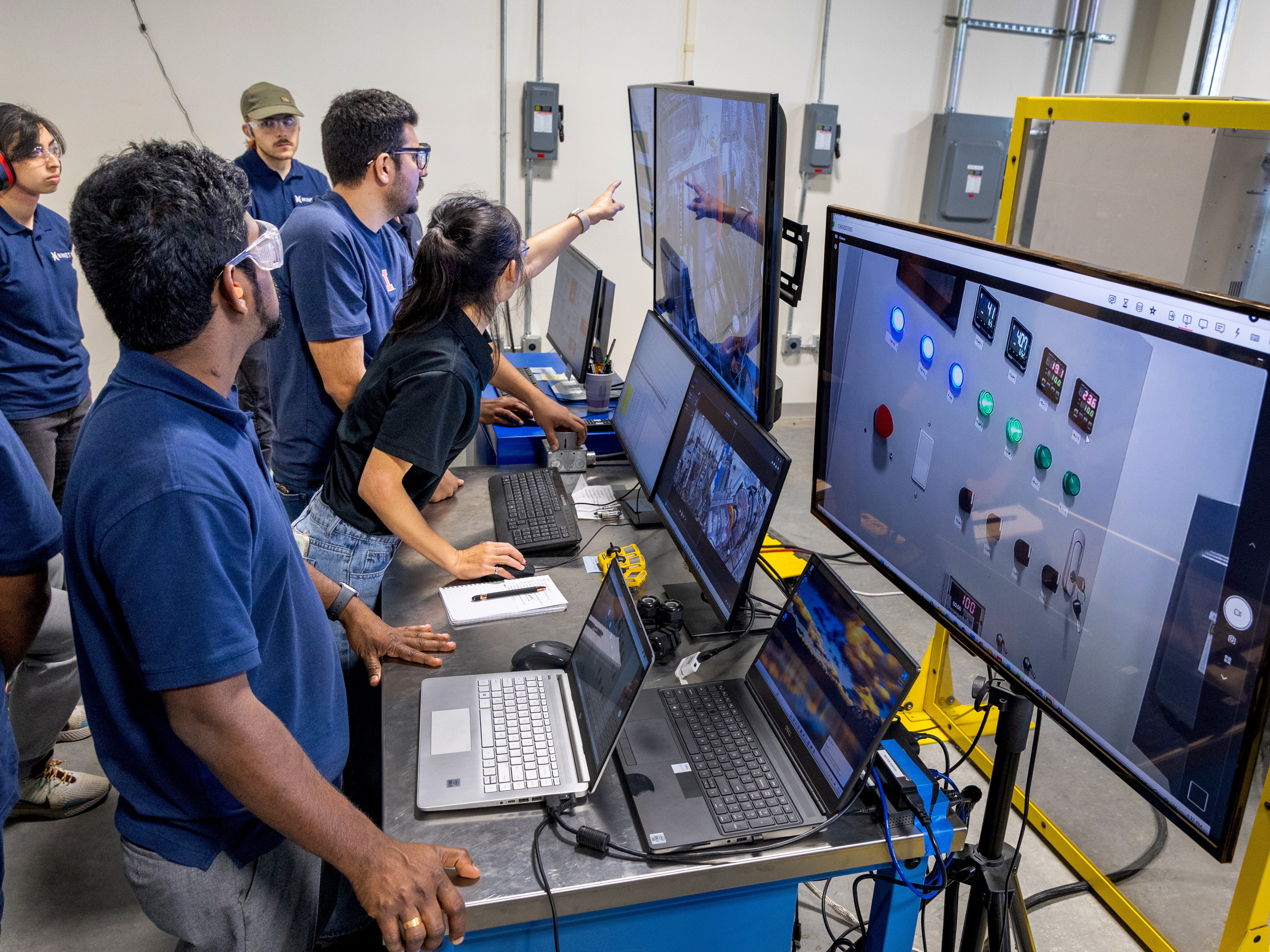 group of students work at laptops and point to several monitors displaying controls and equipment in the POETS Center