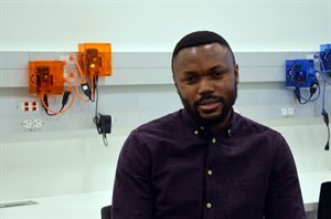 Ajala, pictured with orange and blue distributed control nodes behind him. These devices are geographically dispersed across the power grid. Collectively, they use data acquired via local measurements and via information exchange with other control nodes, to regulate the response of energy resources in the power grid and meet system operational objectives.